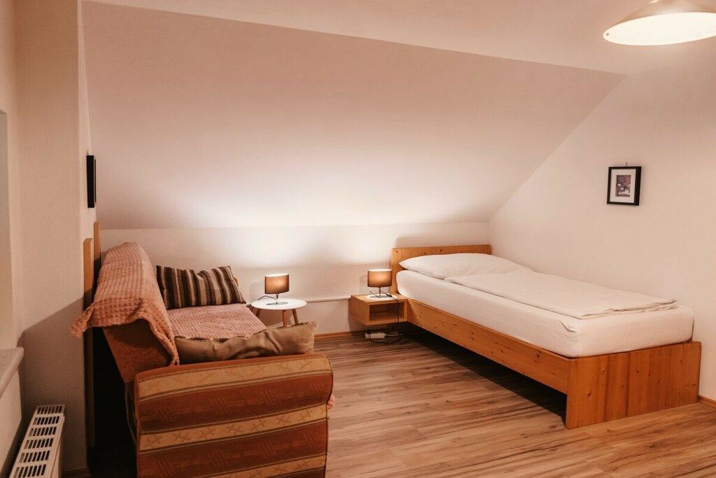 A small, cozy bedroom with a single bed, a wooden nightstand, a bedside table with a lamp, and a cushioned bench.
