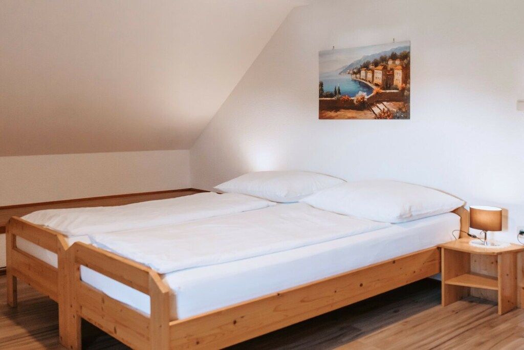 Un lit simple avec un cadre en bois et une literie blanche, dans une pièce dont le plafond est en pente et où l’on peut voir un tableau représentant un paysage côtier.