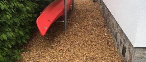 Un espace jardin avec un toboggan rouge et un kayak jaune posé sur un support métallique.