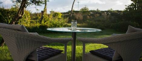 Un espace extérieur aménagé avec des meubles en osier, comprenant une table en verre et des chaises, le tout placé devant un décor de verdure et d’arbres.