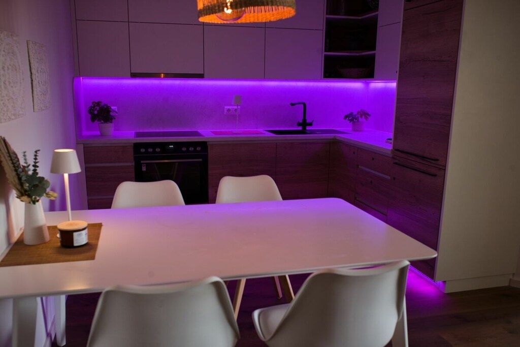 Eine moderne Küche mit Essbereich, ausgestattet mit einem weißen Tisch und Stühlen, einem eingebauten Backofen und einer violett beleuchteten Arbeitsfläche.
