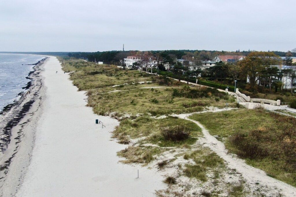 En strand med en sandstig som leder till en gräsyta, med utsikt över ett bostadsområde med hus och en vattenyta.