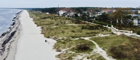 En strand med en sandstig som leder till en gräsyta, med utsikt över ett bostadsområde med hus och en vattenyta.