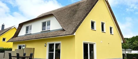 Una casa amarilla con techo de paja, ventanas blancas y un patio con mesa y sillas.