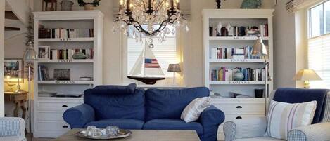 A living room with a blue sofa, a matching armchair, a coffee table, and bookshelves labeled 'ARTS' and 'ROMANS'.