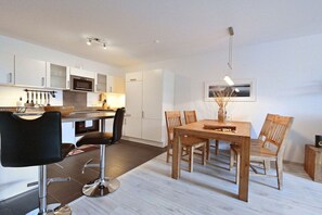 A modern kitchen with white cabinets, a dining area with a wooden table and chairs, and a bar stool. - Holiday apartment with 1 bedroom (Breege\/ Juliusruh OT Juliusruh)