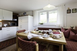 Un comedor con una mesa de madera dispuesta para una comida, una cocina al fondo y una ventana con cortinas.