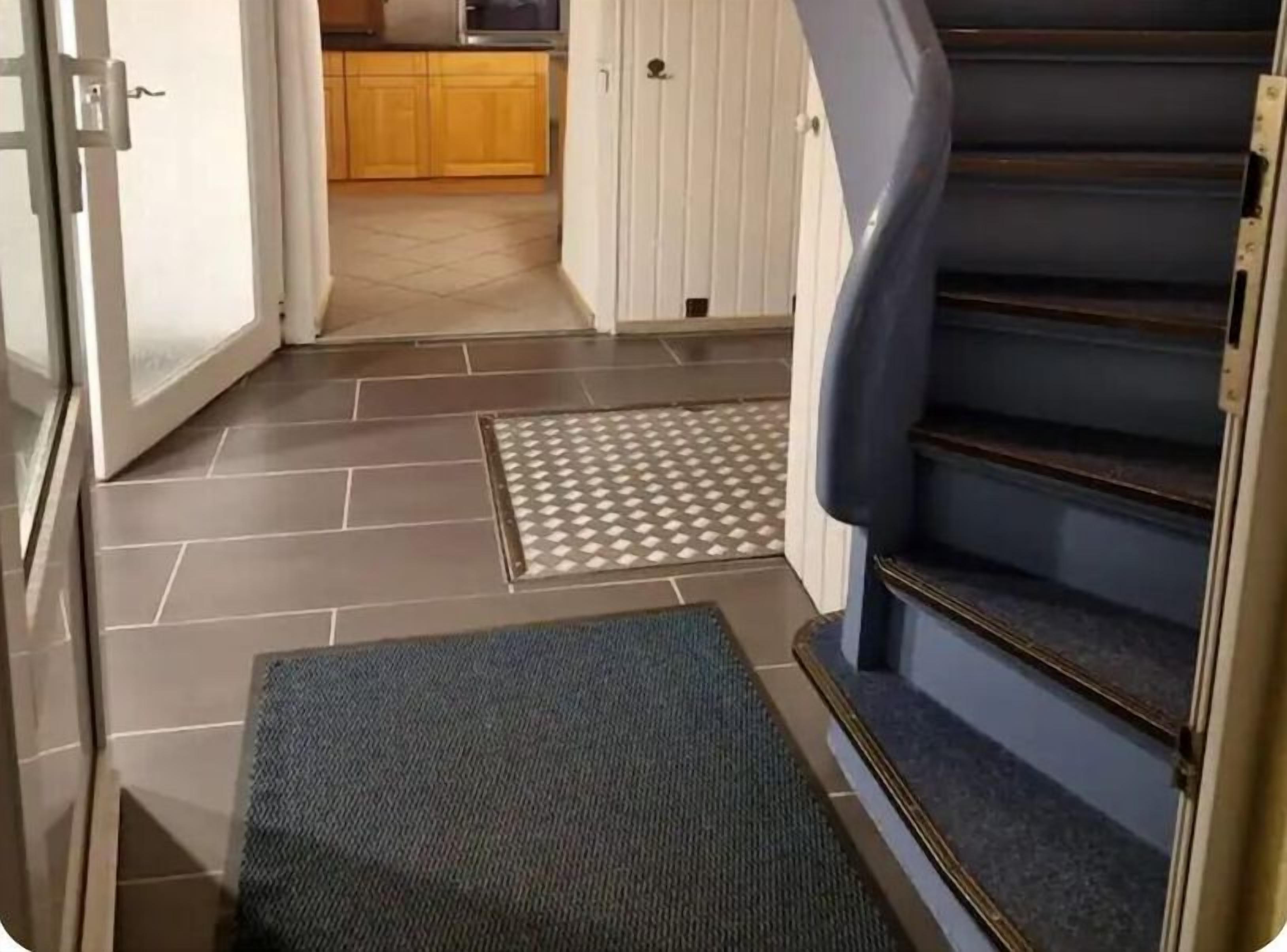 Una escalera conduce a una zona de cocina con piso de baldosas y un felpudo al final.