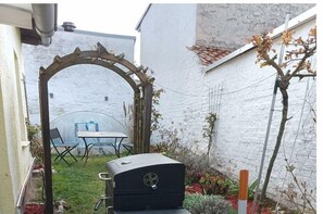 Un patio trasero con una parrilla, un arco de madera, una mesa con sillas y un árbol pequeño.
