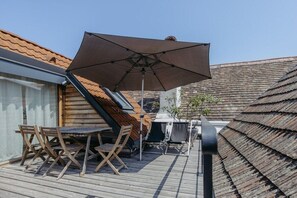 Eine Holzterrasse mit Tisch und Stühlen, einem großen Sonnenschirm und einem Gebäude mit geneigtem Dach.