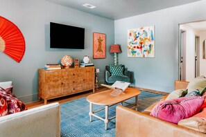 Una sala de estar moderna con un televisor de pantalla plana, un mueble de madera, un tapete azul estampado y un sofá con una manta rosa.