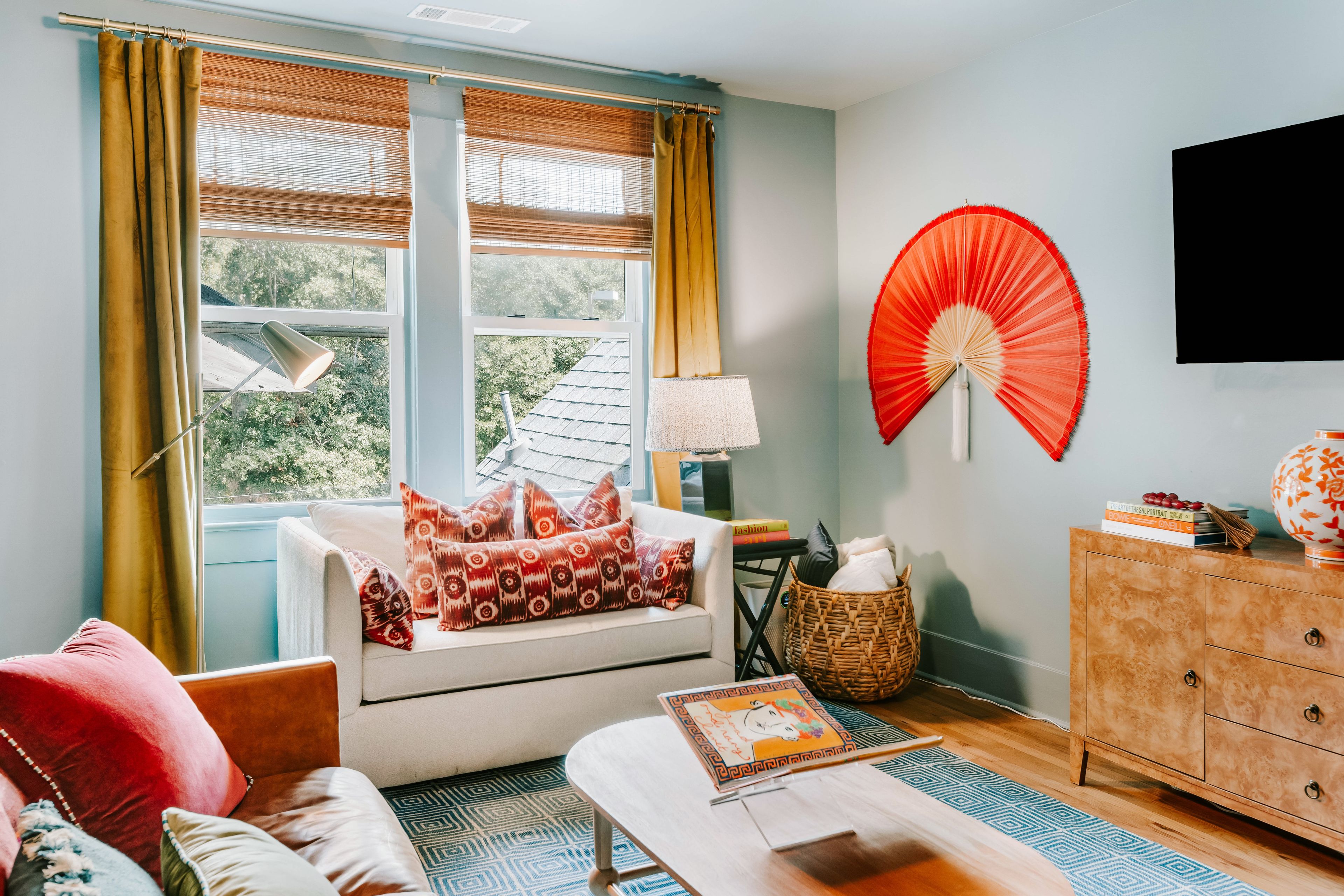 A room with a sofa, a table, a lamp, a fan, and a television on the wall.