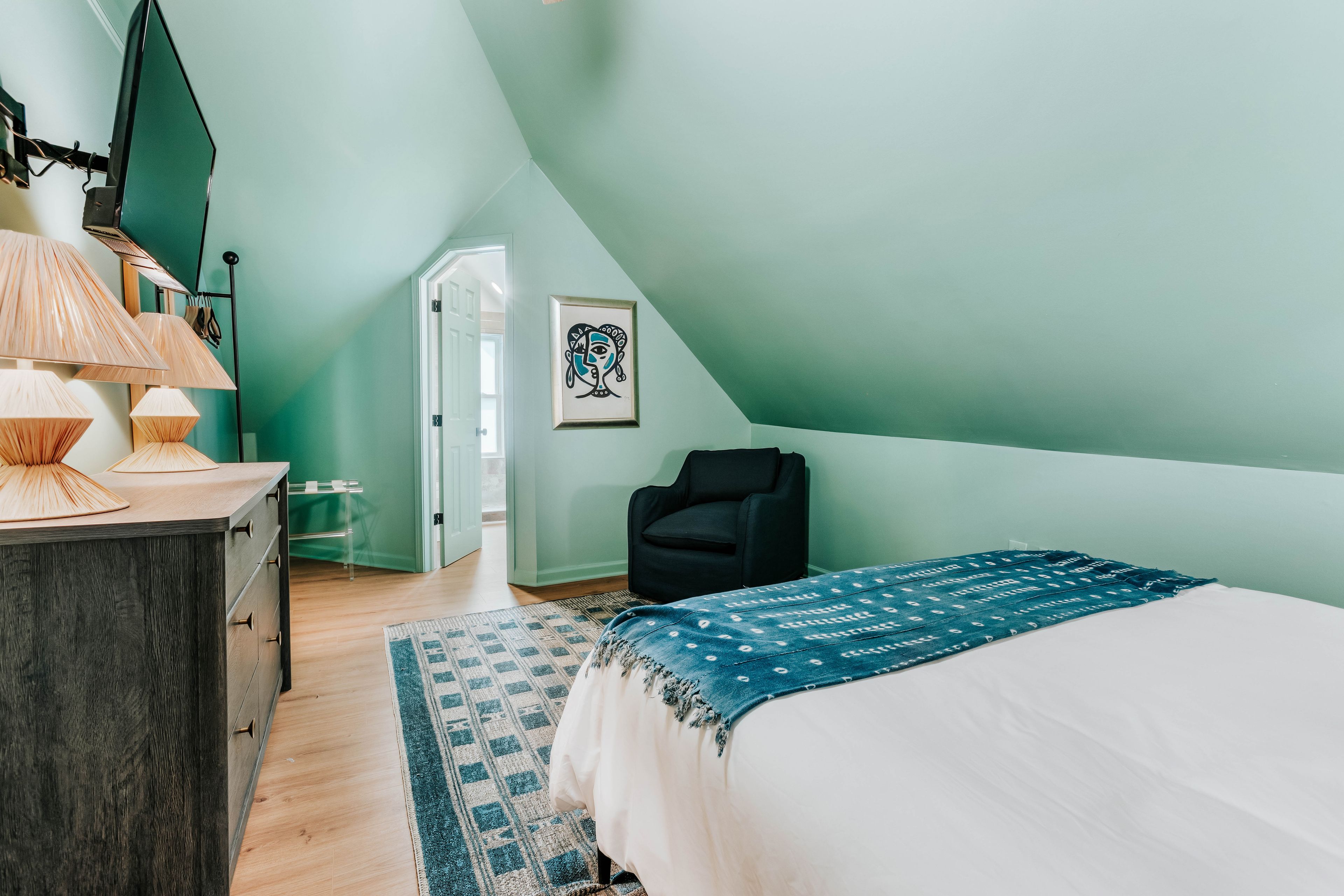 A bedroom with a bed, a chair, a desk with a lamp, and a door leading to another room.
