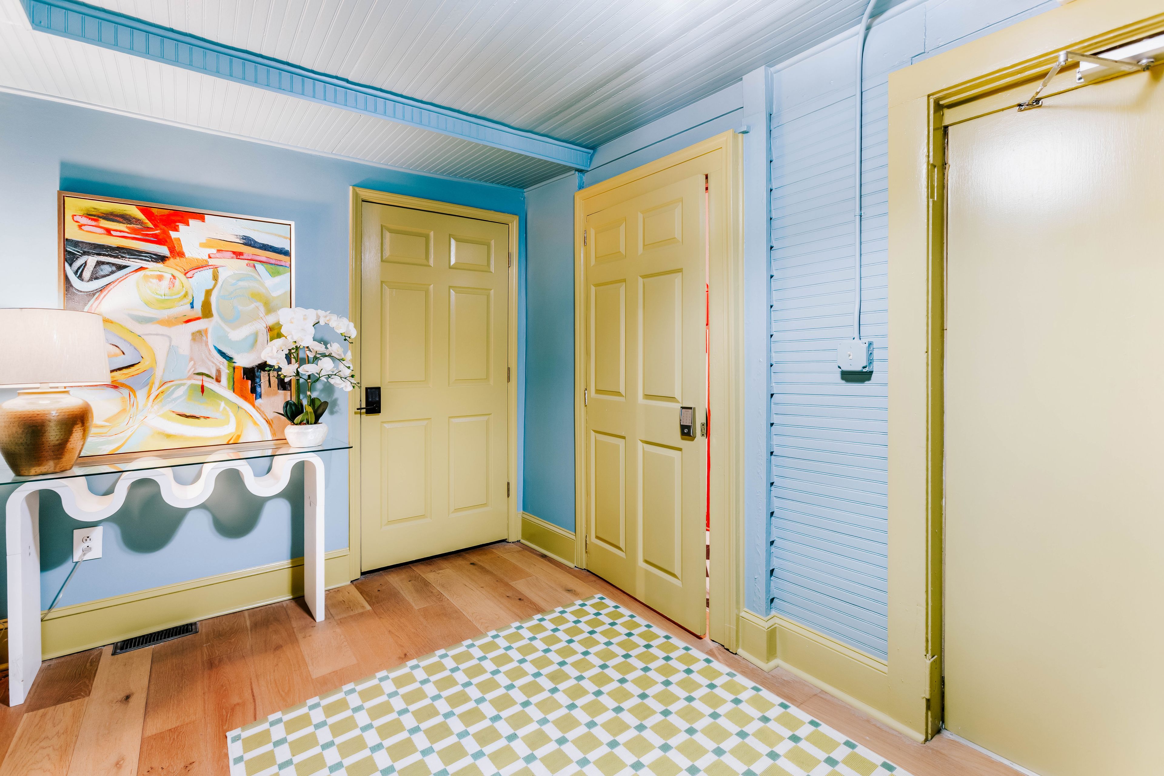 A hallway with a checkered rug, a desk with a vase and a plant, and a door with a keypad lock.