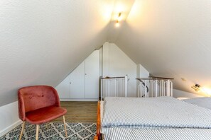 Ein Schlafzimmer mit schrägverdeckter Decke, einem Bett mit gestreifter Bettwäsche, einem roten Sessel und einem gemusterten Teppich.
