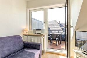 Ein Zimmer mit einer Couch, einem Schreibtisch mit Computer, einem Fernseher und einer Schiebetür zu einer Terrasse mit einem Stuhl.