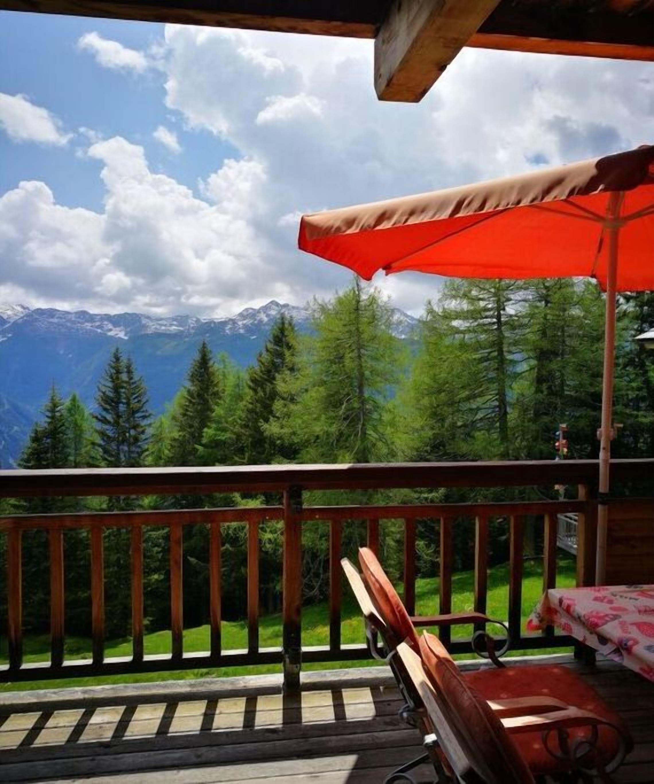 A wooden deck with a red umbrella, a bench, and a table with a floral tablecloth.