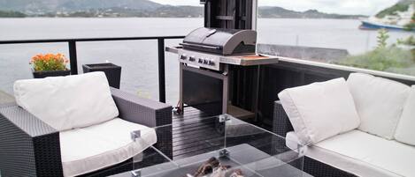 Una terraza al aire libre con mobiliario de mimbre, una parrilla y una mesa de vidrio para hacer fuego.