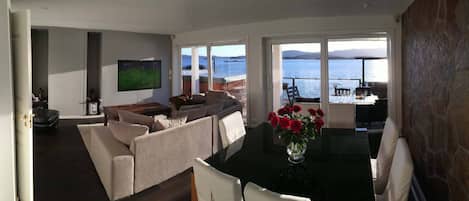 A living room with a sofa, a coffee table, and a TV. There is a balcony with a view of the sea.