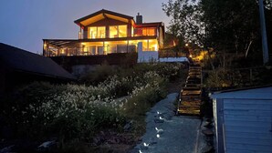 Uma casa moderna de dois andares com fachada de vidro, uma escadaria visível que leva ao andar superior e um caminho com pedras.