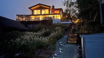 Uma casa moderna de dois andares com fachada de vidro, uma escadaria visível que leva ao andar superior e um caminho com pedras.