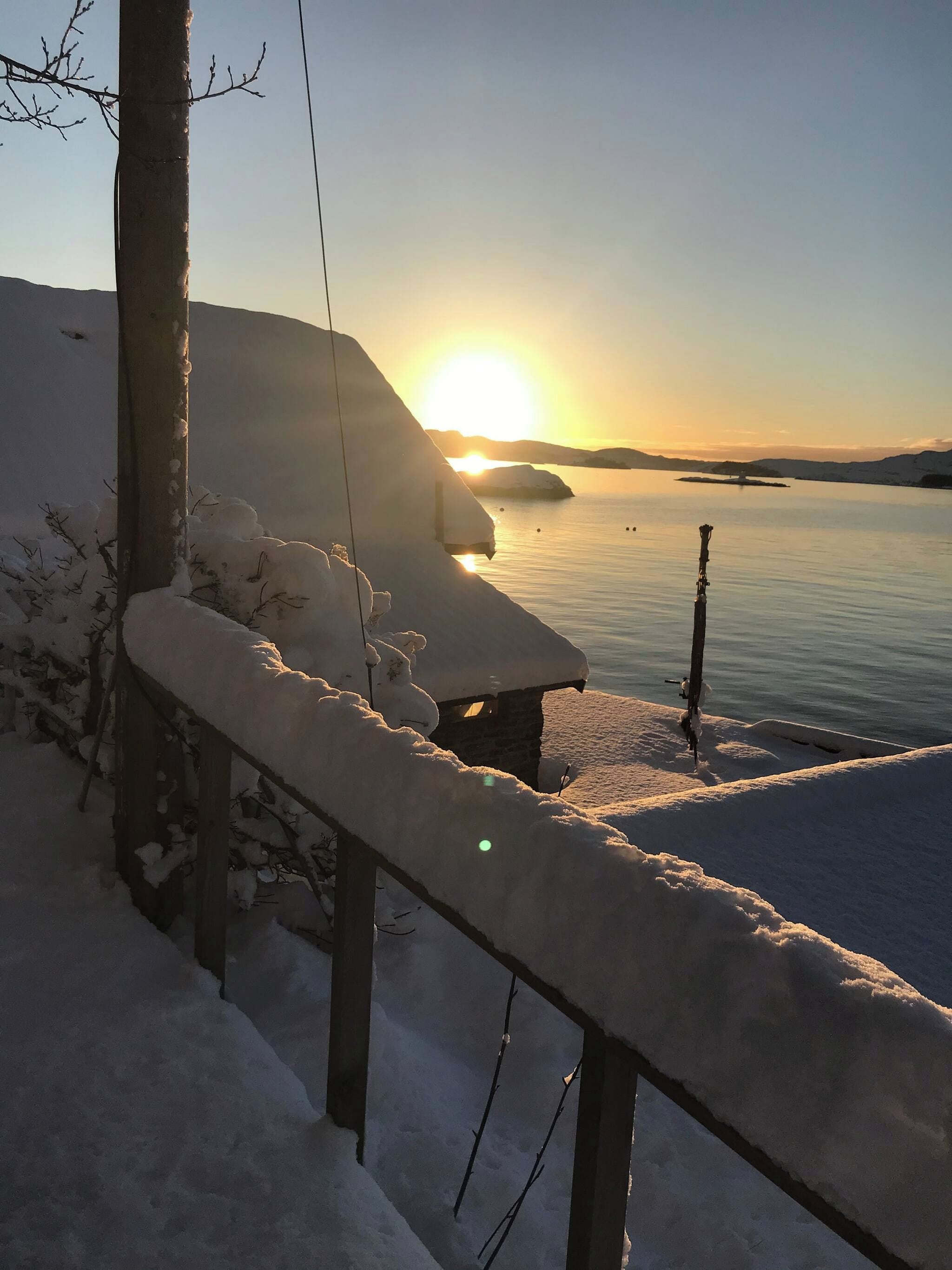 A snow-covered balcony with a view of a calm lake and distant mountains during sunset.