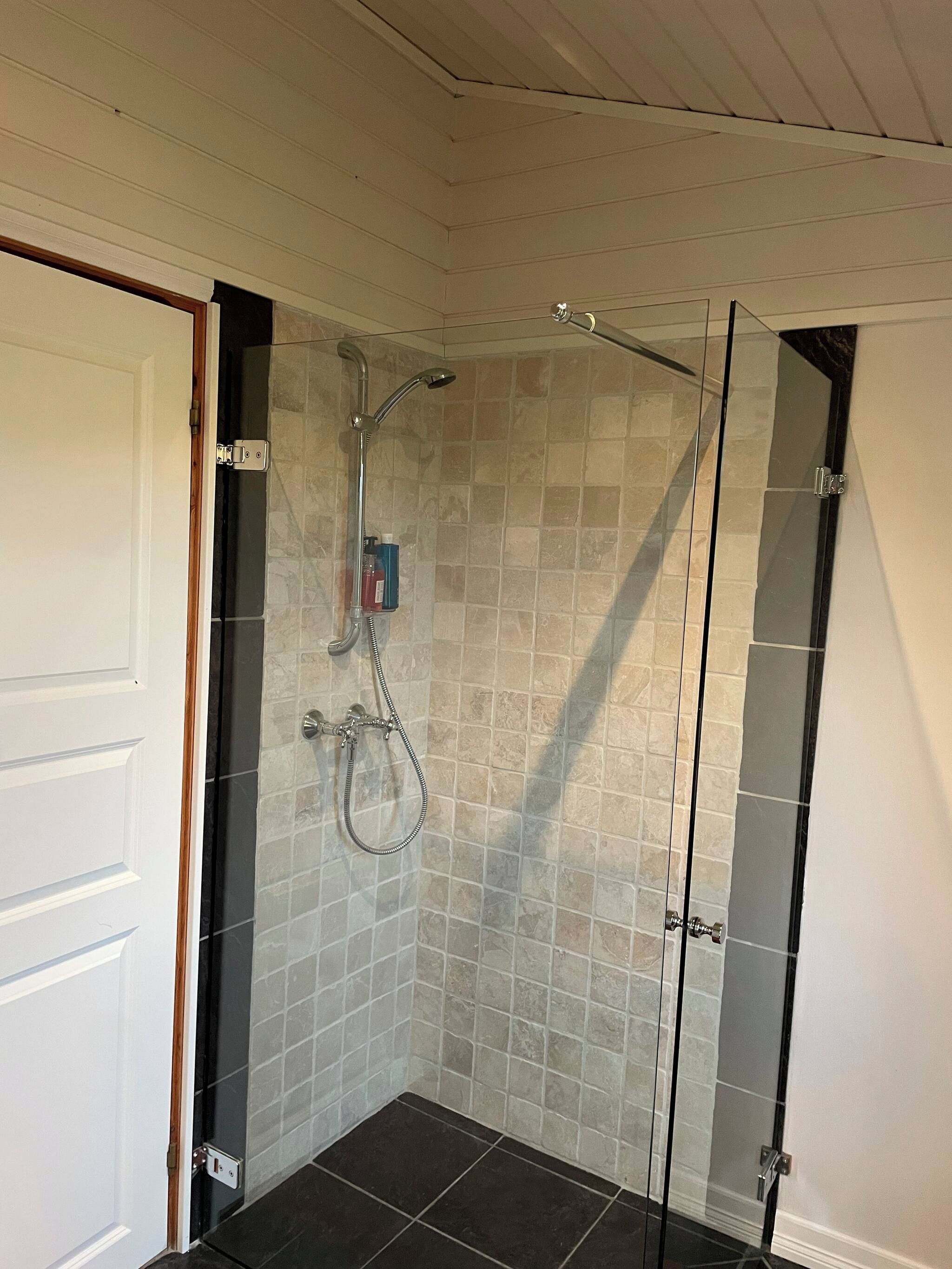 A glass-enclosed shower with a handheld showerhead and a wall-mounted faucet, set against a tiled wall with a door on one side.