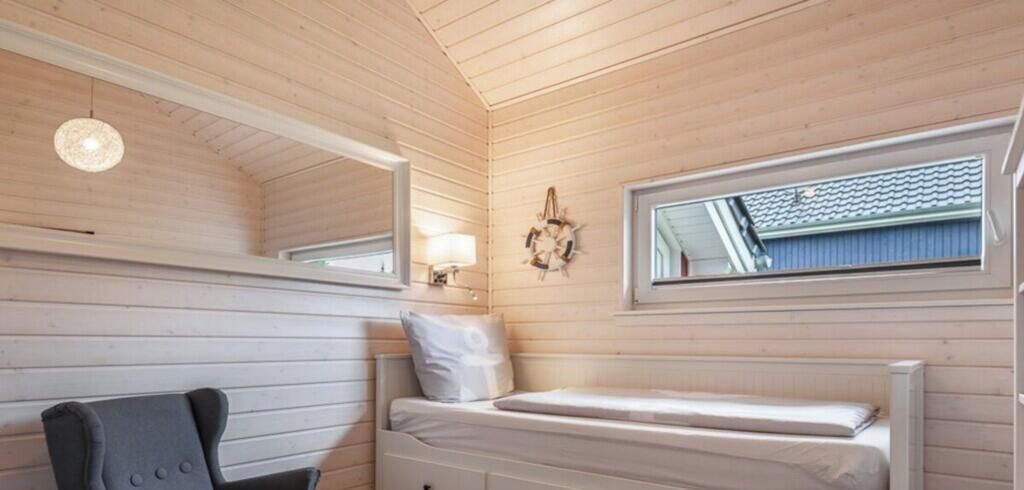 Una cabaña pequeña y con una sola habitación, con cama, sillón, ventana y lámpara de pared.