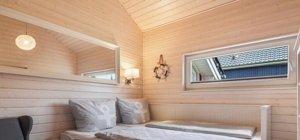 A wooden cabin room with a bed, a chair, a window, and a wall-mounted light fixture.