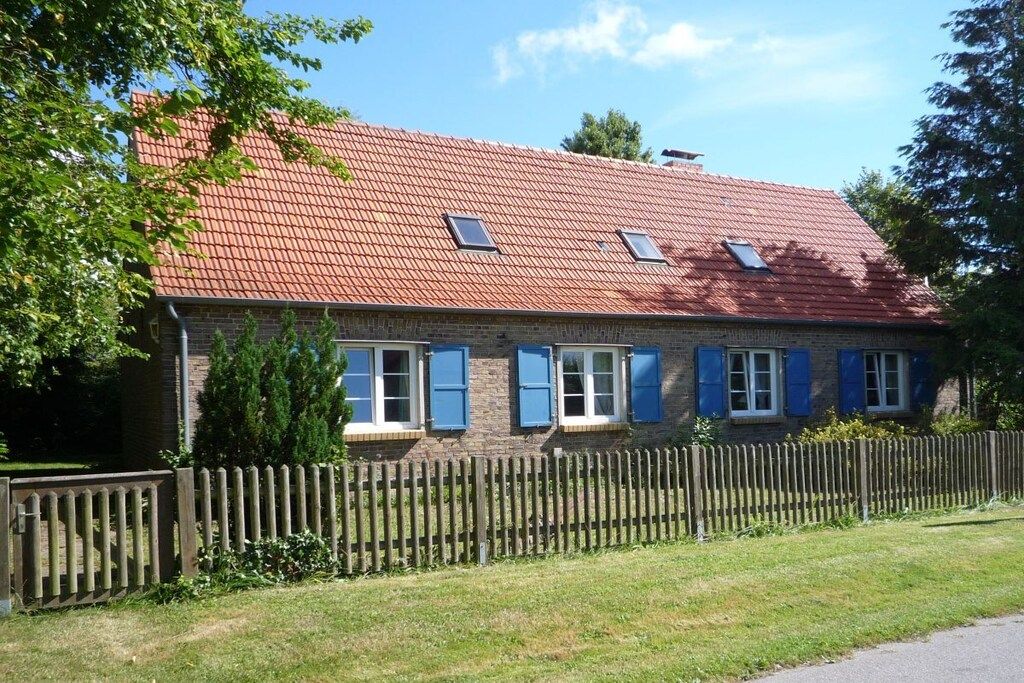 Une maison à deux étages, construite en briques, avec un toit recouvert de tuiles rouges, de nombreuses fenêtres dotées de volets bleus et une clôture en bois à l’avant.