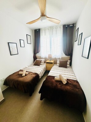 Une chambre avec deux lits, un ventilateur de plafond, une table de chevet et des tableaux encadrés aux murs.