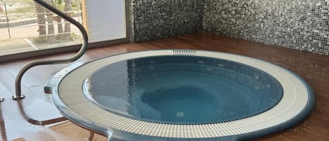 Un jacuzzi con borde texturizado, una escalera metálica y una pared de azulejos con diseño tipo mosaico.