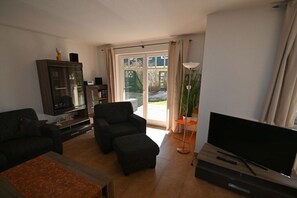 Un salon comprenant un canapé, un fauteuil, un pouf, une télévision et un meuble.