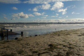 A beach with a wooden pier extending into the water, waves crashing onto the shore, and a clear sky with scattered clouds. - LEE in the Captain's House Ostseerose (Travemünde)