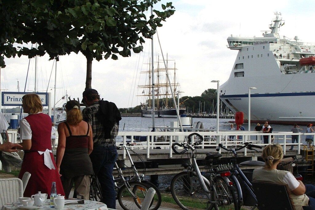 A waterfront scene with a large ship docked, people standing and sitting, and bicycles parked.