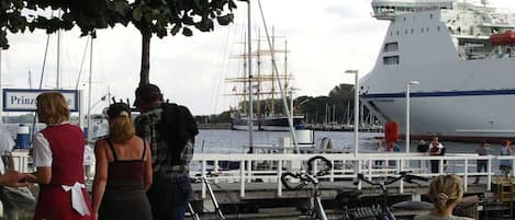 Eine Uferansicht mit einem großen Schiff, das vertäut ist, Personen, die stehen und sitzen, sowie abgestellten Fahrrädern.