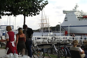 A waterfront scene with a large ship docked, people standing and sitting, and bicycles parked. - LEE in the Captain's House Ostseerose (Travemünde)