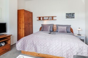 A bedroom with a bed, wooden wardrobe, bedside table, lamp, and a shelf with items. - LEE in the Captain's House Ostseerose (Travemünde)