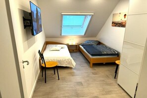 Quarto pequeno e bem iluminado com teto inclinado, uma cama de solteiro, uma cadeira, uma mesa e uma TV na parede.
