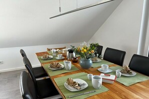 Uma área de refeições com mesa de madeira posta para quatro pessoas, incluindo pratos, copos e um vaso com flores.