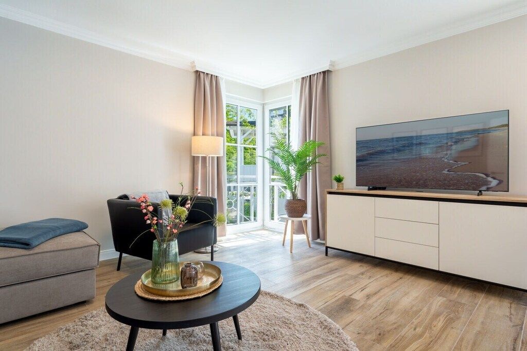 Una sala moderna con un sofá, una mesa de centro con un jarrón, un televisor sobre un mueble bajo y un ventanal con cortinas.