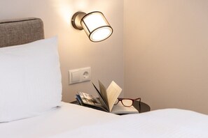 Ein ordentlich gemachtes Bett mit einem weißen Kopfkissen, ein Nachttisch mit einem Buch und einer Brille sowie eine Wandlampe.