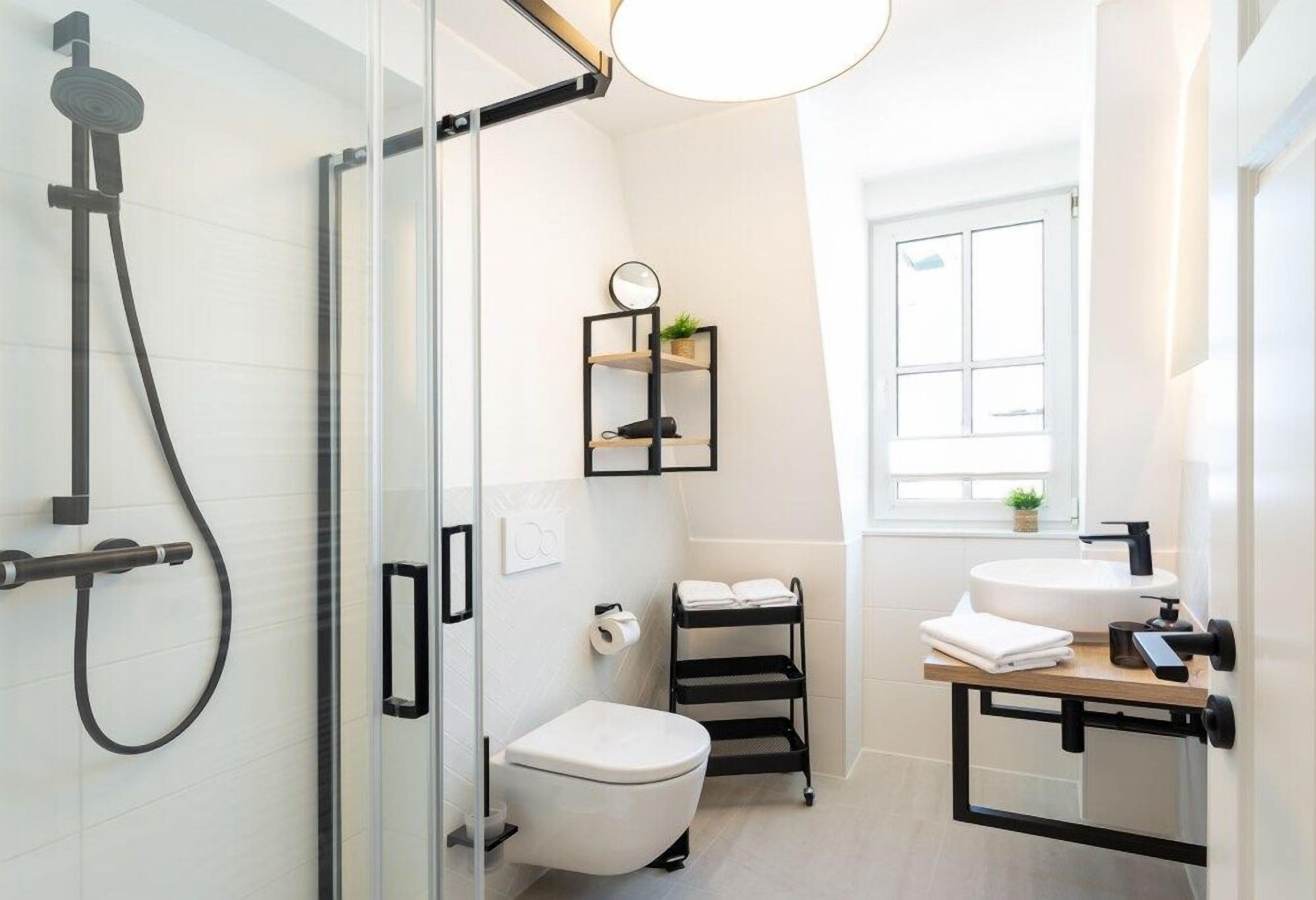 Un baño moderno con cabina de ducha de vidrio, inodoro, lavamanos con grifo negro y una pequeña ventana que deja entrar luz natural.