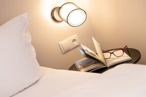 Una cama con un almohadón, una mesita de noche con un libro y gafas, y una lámpara fijada a la pared.