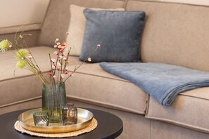 Una sala de estar con un sofá, un cojín azul, un florero de cristal con flores y una bandeja con cristalería.