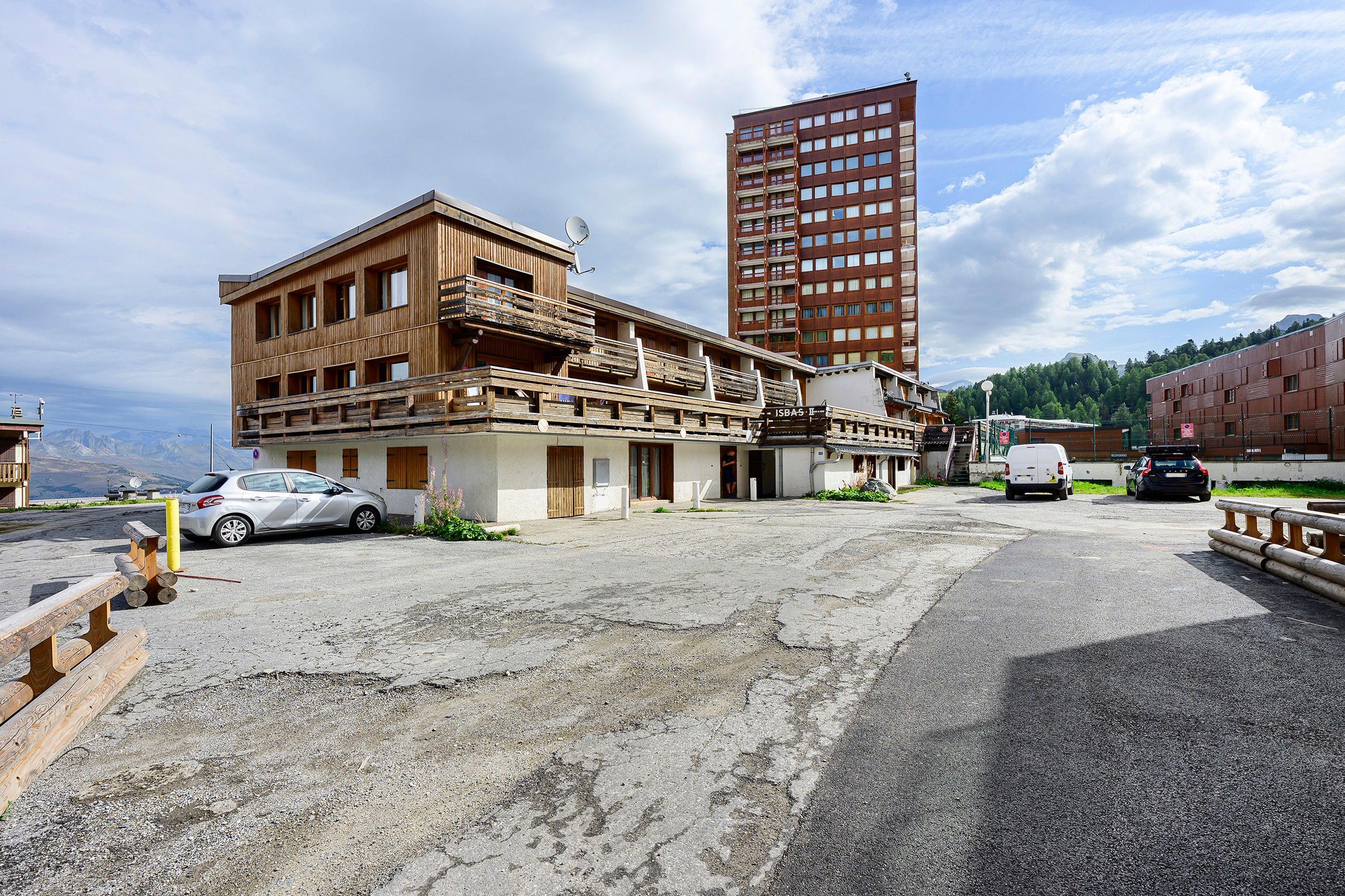 Un bâtiment à la façade en bois et doté de balcons, un espace de stationnement avec des voitures, et un grand immeuble rouge en arrière-plan.
