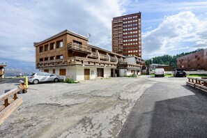 Un edificio con fachada de madera y balcones, un estacionamiento con coches, y un edificio alto de color rojo al fondo.