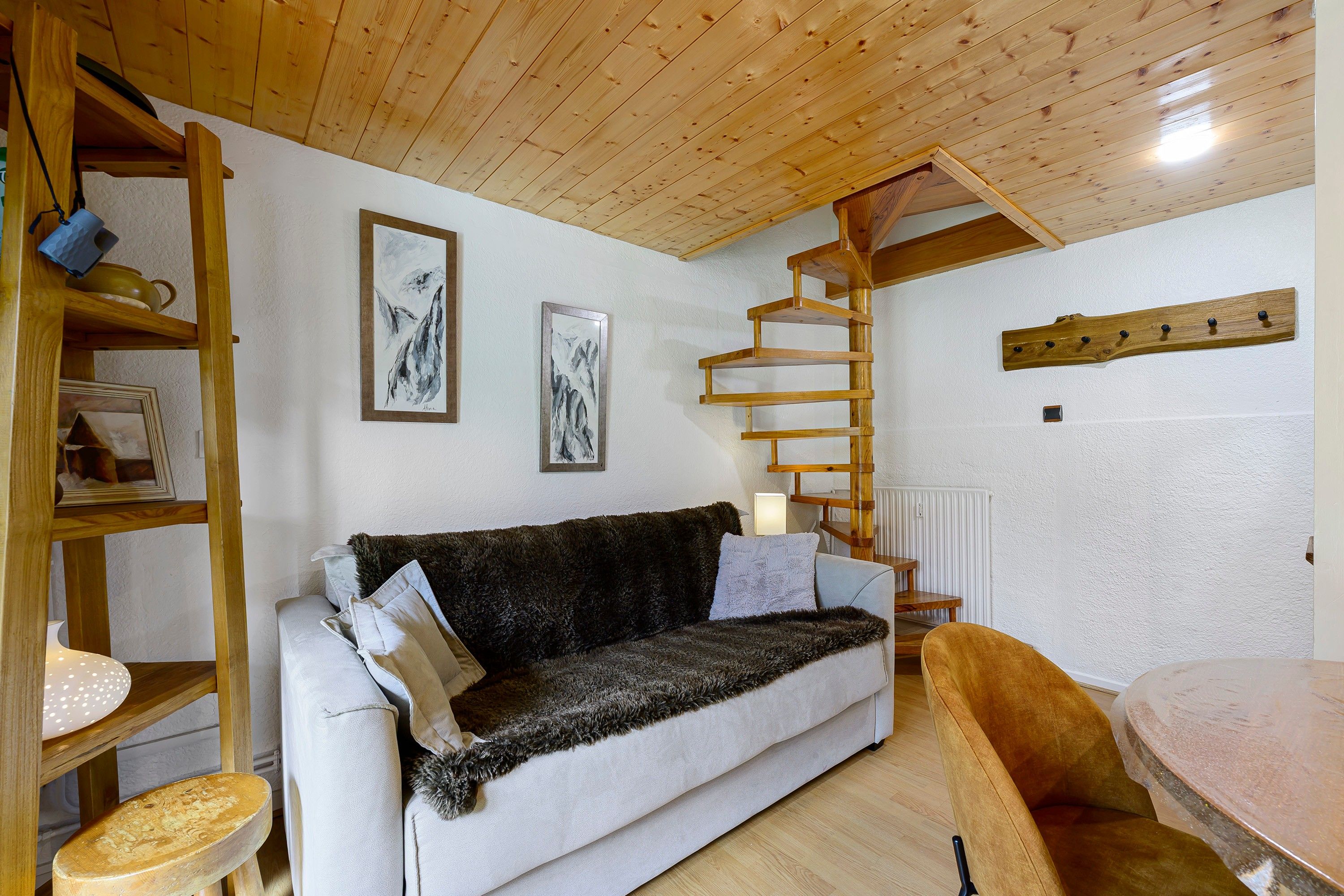 Une pièce chaleureuse avec un plafond en bois, un canapé blanc, une échelle en bois et deux œuvres d’art encadrées accrochées au mur.