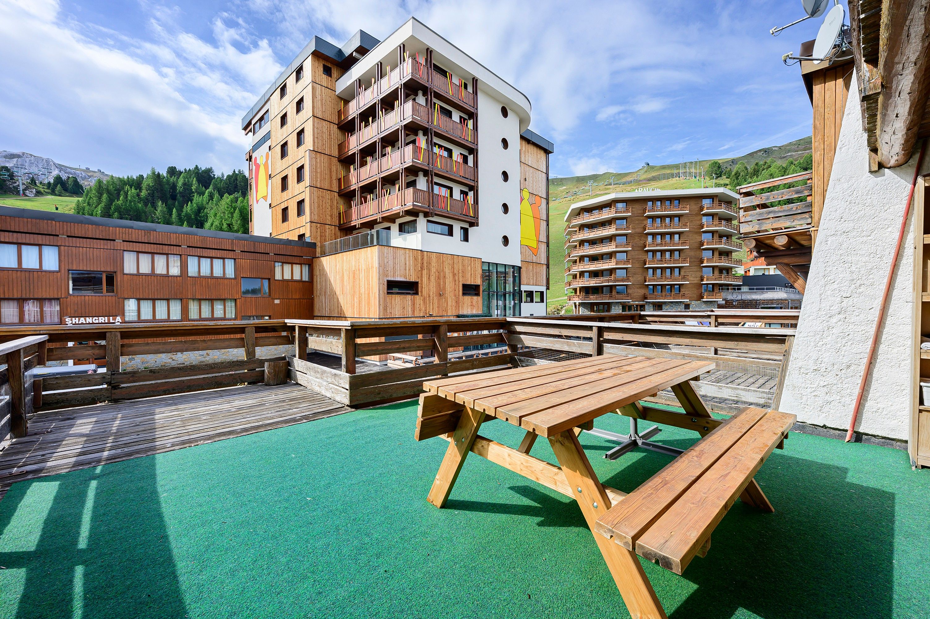 A wooden deck with picnic tables, a building with multiple balconies, and a mountainous backdrop.