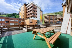 Una terraza de madera con mesas de picnic, un edificio con varios balcones y un telón de fondo montañoso.